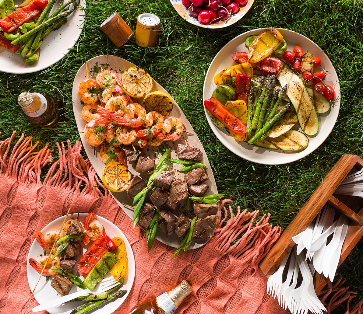 Plates of grilled vegetables, meat, and seafood laid out on the grass